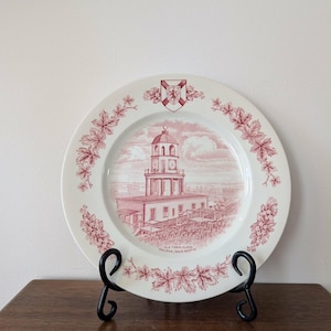 May include: A white decorative plate with a red illustration of a building with a clock tower, surrounded by a floral border. The plate has the text "OLD TOWN CLOCK HALIFAX, NOVA SCOTIA". The plate is displayed on a black metal stand.