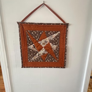 May include: A square, quilted wall hanging featuring a star design. The star is crafted from brown and cream patterned fabric, set against a rust-coloured background. It has a rust border and a brown leather hanging strap.
