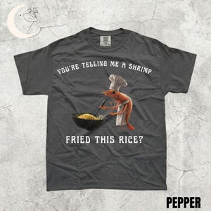 May include: Dark grey t-shirt featuring the text "YOU'RE TELLING ME A SHRIMP FRIED THIS RICE?" and a cartoon shrimp in a chef's hat and apron, cooking rice in a wok. The word "PEPPER" is at the bottom of the shirt.