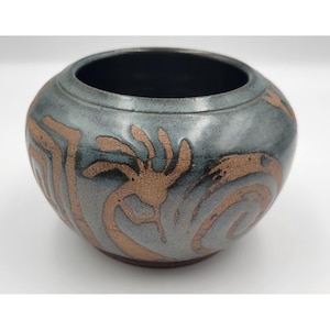 May include: A round ceramic bowl with a metallic charcoal gray glaze and a wide opening. The exterior features textured, copper-colored etched designs of a flute player and swirling patterns that contrast against the dark, glossy surface.