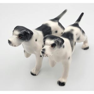 Puede incluir: Dos figuras de perros de cerámica. Los perros son blancos con manchas negras y orejas negras. Están posados en una postura de caminar, con las colas levantadas. Las figuras tienen ojos marrones y están sobre un fondo blanco.