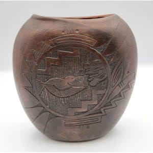 May include: A dark brown, rounded clay vase featuring an etched design of a hummingbird hovering near a flower. The surface displays geometric patterns, including zig-zag lines and circular motifs, carved into the matte, earthy-toned pottery.