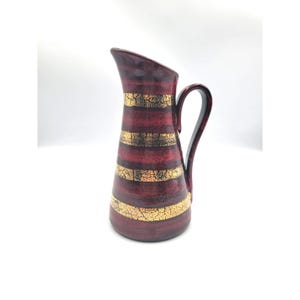 May include: A maroon ceramic pitcher with a gold crackle stripe design. The pitcher has a curved handle and a slanted spout. The gold stripes are horizontal and evenly spaced around the body of the pitcher.