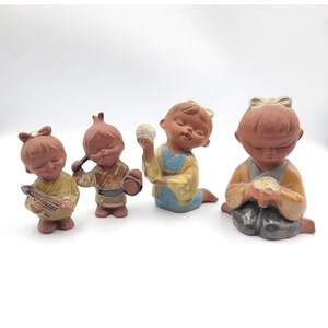 May include: Four terracotta figurines of children in traditional attire. One plays a stringed instrument, another holds a drum and mallet, a third holds a ball, and the last one is seated with hands clasped. Each figure has closed eyes.