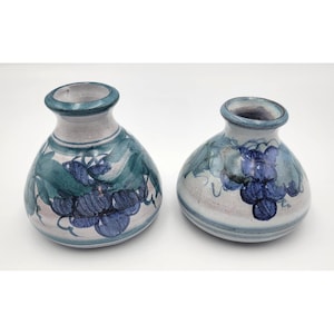 May include: Two ceramic vases with a rounded base and a narrow neck. The vases feature a hand-painted design of blue grapes and green leaves on a light gray background, with blue bands around the rim and base of each piece.