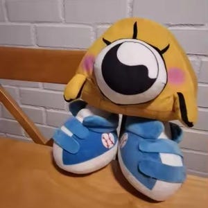 May include: A plush toy featuring a yellow, eye-shaped head with a large black and white eye, pink cheeks, and black eyelashes. It is paired with blue sneakers with white accents and baseball details. The toy is set on a wooden surface.