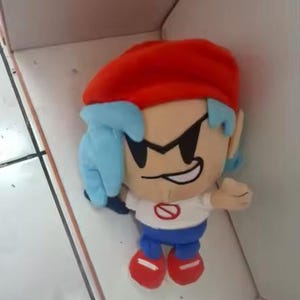 May include: A plush doll of a cartoon character with a red hat, blue hair, and a white shirt with a red circle. The doll has black sunglasses and blue pants with red shoes. The doll is sitting in a white box.