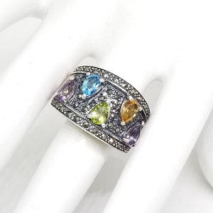 May include: A wide silver band ring featuring a textured, dark oxidized surface embedded with small sparkling stones. The ring is set with five pear-shaped faceted gemstones in shades of light blue, light green, yellow, and purple.