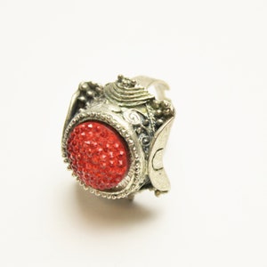 May include: Silver-toned ring with a large, oval, red, jeweled centerpiece. The ring has ornate detailing around the red jewel and on the band. The ring is set against a white background.