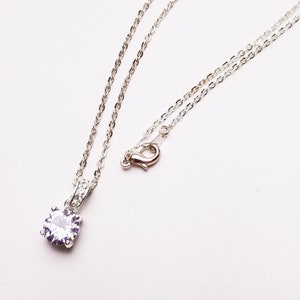 May include: A silver necklace with a delicate chain and a single, round, light purple gemstone pendant. The pendant is set in a silver setting and is the focal point of the necklace. The chain has a lobster clasp.