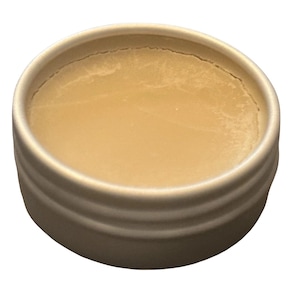 May include: A small, round, metal container filled with a light beige balm. The container has a ridged design around the sides. The balm has a smooth, slightly textured surface.