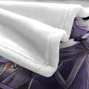 May include: A white plush blanket featuring a character from The Nightmare Before Christmas. The blanket has a soft, textured surface and a detailed image of Jack Skellington in a pinstripe suit. The blanket's edge is neatly finished.