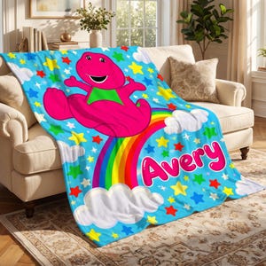 May include: A bright blue fleece blanket featuring a large pink dinosaur character with a green belly, a colorful rainbow, white clouds, and scattered multi-colored stars. The name Avery is printed in large, bold pink letters on the blanket.
