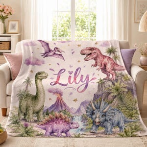 May include: A soft, cream-colored fleece blanket featuring a colorful watercolor print of various dinosaurs, including a green long-neck, a pink T-Rex, a purple stegosaurus, and a blue triceratops, with the name Lily printed in purple script.