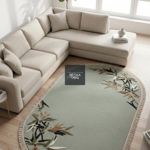 May include: Oval sage green rug with a fringed edge. The rug has a botanical design with brown, black, and white leaves and flowers. It is placed on a wooden floor in a living room setting, near a beige sofa.