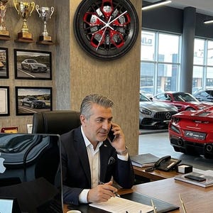 Puede incluir: Un reloj de pared circular diseñado para parecerse a una rueda de coche negra y roja con pinzas de freno y manecillas blancas. El reloj presenta números romanos en el borde exterior y está montado en una pared sobre un escritorio de oficina.