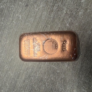 May include: A copper bar, wrapped in clear plastic, with the text "GEIGER EDELMETALLE" and "FEINKUPFER 999.9" visible. A coin is placed on top of the bar. The bar is marked with "500g".