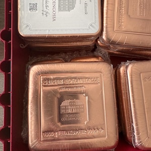May include: A collection of copper bars, each wrapped in clear plastic. The bars feature the text "Geiger Edelmetalle" and "Schloss Güldengossa". The bars are marked as 1000g Feinkupfer 999.9.
