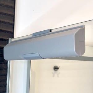 May include: A light gray shower door header with a rectangular design. The header is mounted above a glass shower door. The shower door is in a white bathroom.