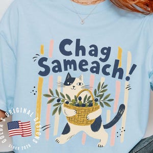 May include: A light blue t-shirt featuring a graphic of a white and black cat carrying a woven basket filled with olive branches. Above the cat, the text reads "Chag Sameach!" in dark blue letters. Vertical pastel stripes appear behind the cat.