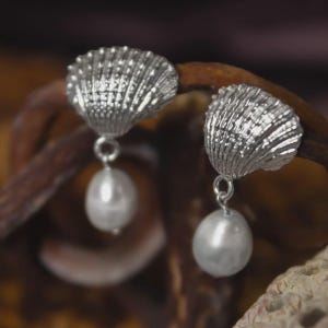 Puede incluir: Un par de pendientes de concha plateados con perlas blancas colgantes en forma de pera. Los pendientes presentan diseños detallados de conchas y están suspendidos de pequeños bucles. El fondo es una mezcla borrosa de marrón y beige.