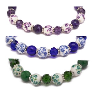May include: Three bracelets with floral patterned beads and crystal accents. The first bracelet has purple beads, the second has blue beads, and the third has green beads.