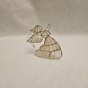 May include: A decorative angel ornament crafted from thin gold-toned wire and translucent white panels. The figure features delicate wire-work wings and a flowing gown design with intricate swirling patterns integrated into the metal frame structure.