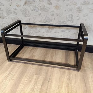 Könnte beinhalten: Ein rechteckiger Schuhständer mit offenem Rahmen aus dunklem Metall. Der Ständer hat ein schlichtes, modernes Design mit einem horizontalen Regal und abgewinkelten Beinen. Der Ständer steht auf einem hellen Holzboden.