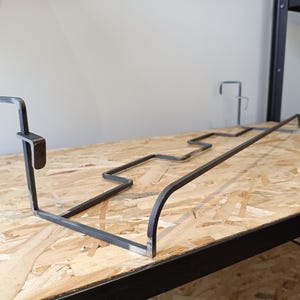May include: A dark gray metal rack featuring a stepped, zig-zag design with two integrated hooks on the sides for mounting. The structure is made of bent metal rods, providing a functional frame for organizing or displaying items in a workshop.