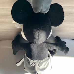 May include: A plush toy of a black and white cartoon character with large black ears, a gray hat, and gray shorts. The toy has black arms and a black tail. The character's face is white with black eyes.