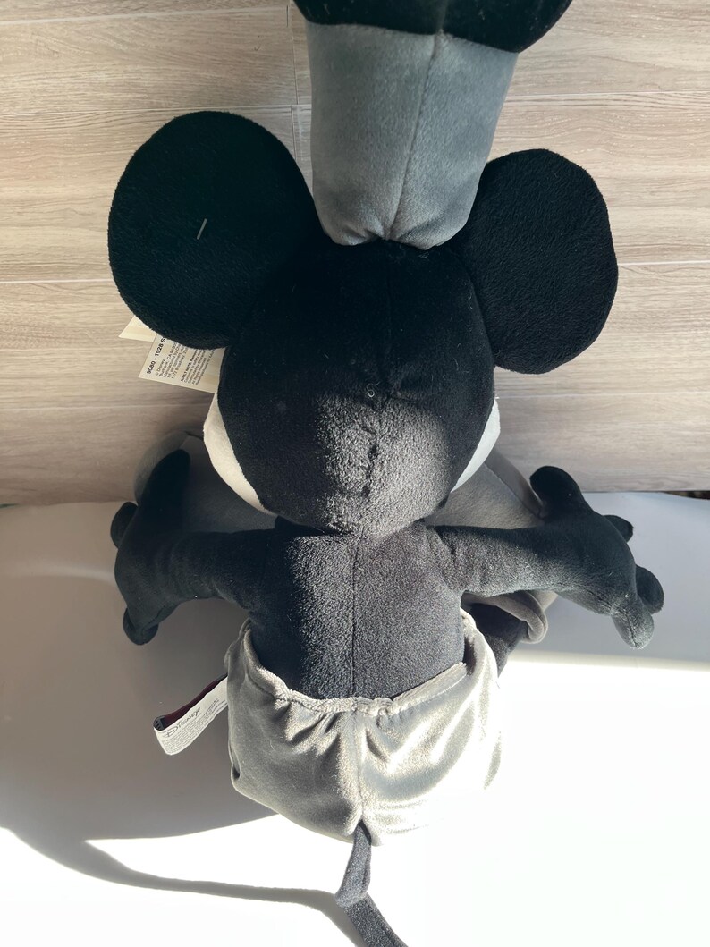May include: A plush toy of a black and white cartoon character with large black ears, a gray hat, and gray shorts. The toy has black arms and a black tail. The character's face is white with black eyes.