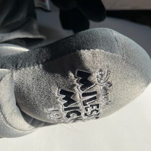 May include: Close-up of a gray plush toy foot with the words "MILESTONE MICKEY" embroidered in black. The plush toy is gray and black, with a soft, velvety texture. The image is well-lit, and the details of the embroidery are clear.