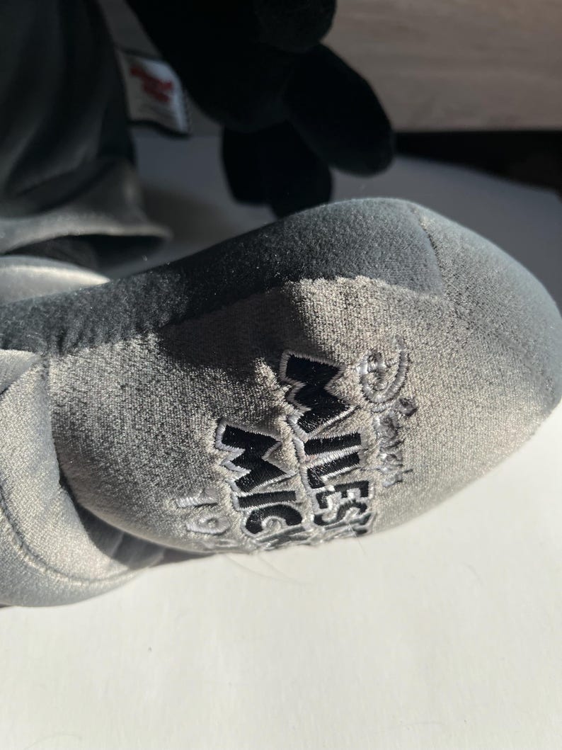 May include: Close-up of a gray plush toy foot with the words "MILESTONE MICKEY" embroidered in black. The plush toy is gray and black, with a soft, velvety texture. The image is well-lit, and the details of the embroidery are clear.