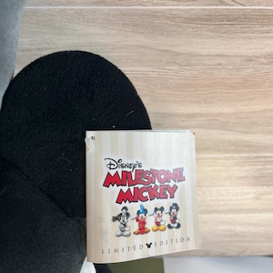 May include: A black and white plush Mickey Mouse toy with a tag that reads "Disney's Milestone Mickey" and "Limited Edition". The tag features four different Mickey Mouse designs. The toy is set against a light wood grain background.