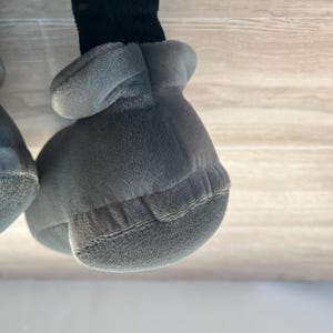 May include: Close-up of a plush toy's gray gloved hand and black leg. The glove is a soft, rounded shape with a textured surface and stitched details. The leg is a dark, fuzzy material.
