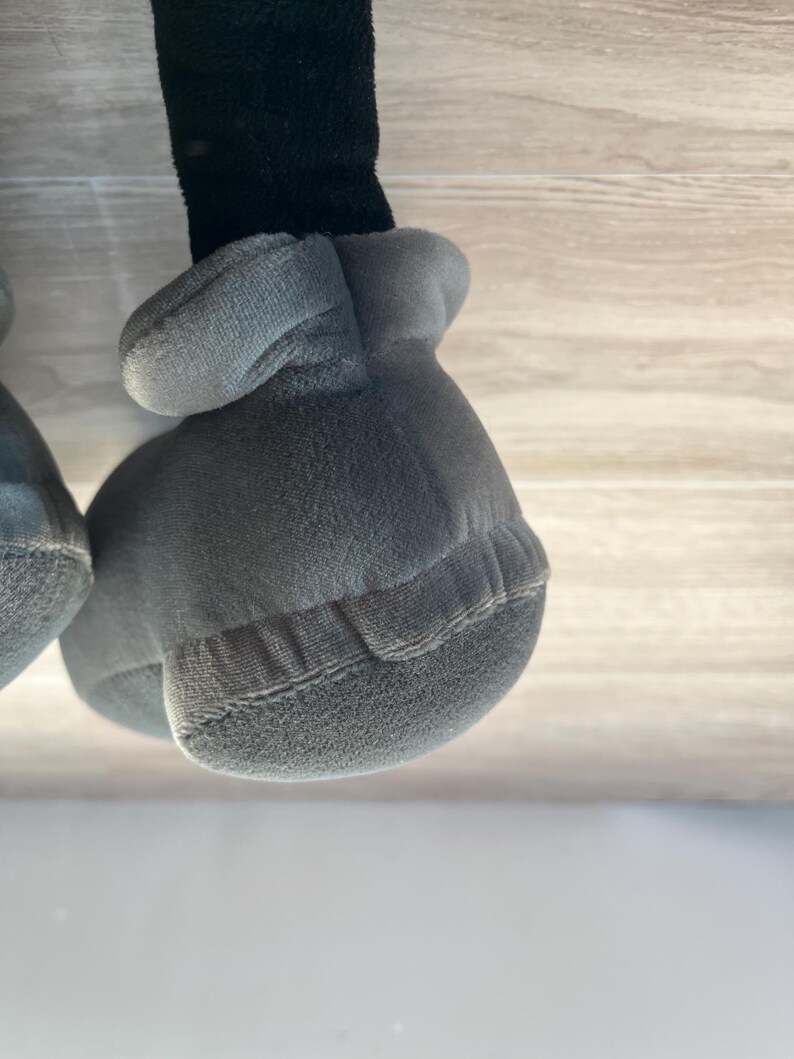 May include: Close-up of a plush toy's gray gloved hand and black leg. The glove is a soft, rounded shape with a textured surface and stitched details. The leg is a dark, fuzzy material.