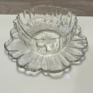 May include: A clear glass bowl and matching plate set featuring a scalloped, petal-like edge design. The bowl has vertical fluted sides and a smooth base, while the plate mirrors the floral shape with wide, curved glass segments throughout.