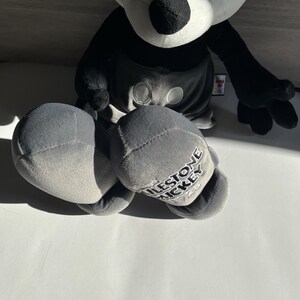May include: A plush Mickey Mouse doll in black, white, and gray. The doll has large black eyes, a white face, and black ears. The gray shorts have two white buttons. The doll's foot reads "MILESTONE MICKEY".