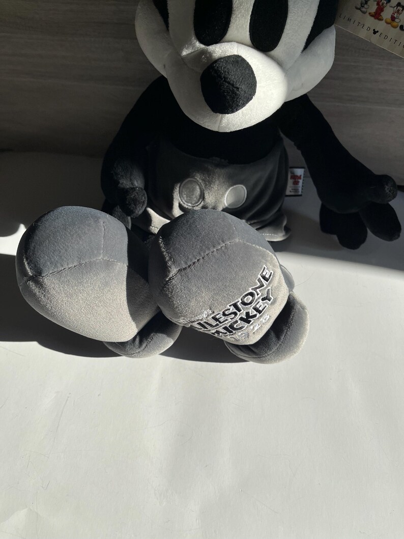 May include: A plush Mickey Mouse doll in black, white, and gray. The doll has large black eyes, a white face, and black ears. The gray shorts have two white buttons. The doll's foot reads "MILESTONE MICKEY".