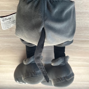 May include: A plush Mickey Mouse toy, viewed from the back, showcasing gray shorts, black legs, and large gray shoes. The tail is visible. The Disney tag is on the left side.