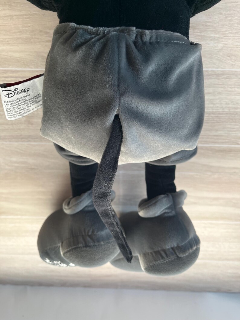 May include: A plush Mickey Mouse toy, viewed from the back, showcasing gray shorts, black legs, and large gray shoes. The tail is visible. The Disney tag is on the left side.
