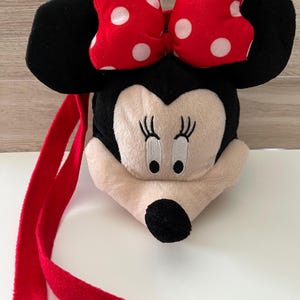 May include: A plush Minnie Mouse-shaped bag with a red strap. The bag features a red bow with white polka dots, black ears, and a cream-colored face with embroidered details. The strap is a vibrant red.