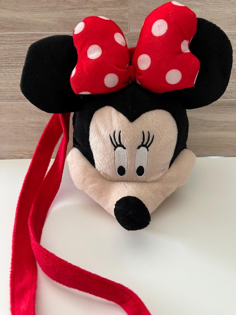 May include: A plush Minnie Mouse-shaped bag with a red strap. The bag features a red bow with white polka dots, black ears, and a cream-colored face with embroidered details. The strap is a vibrant red.