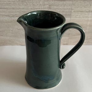 May include: A dark forest green ceramic pitcher with a glossy finish, featuring a wide cylindrical body, a curved handle with a curled base, and a distinct pouring spout at the top rim. The surface shows subtle variations in the deep green glaze.