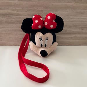 May include: A plush Minnie Mouse-shaped bag with a red strap. The bag features a black head with large black ears, a cream-colored face, and a red bow with white polka dots. The bag is designed to be worn over the shoulder.