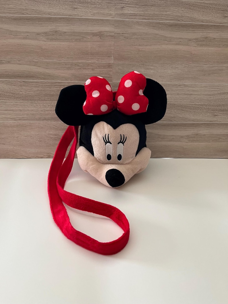 May include: A plush Minnie Mouse-shaped bag with a red strap. The bag features a black head with large black ears, a cream-colored face, and a red bow with white polka dots. The bag is designed to be worn over the shoulder.