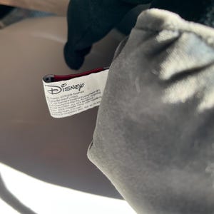 May include: Close-up of a plush toy with a gray body and dark green head. A white tag with the Disney logo and text is visible. The tag is red and white.