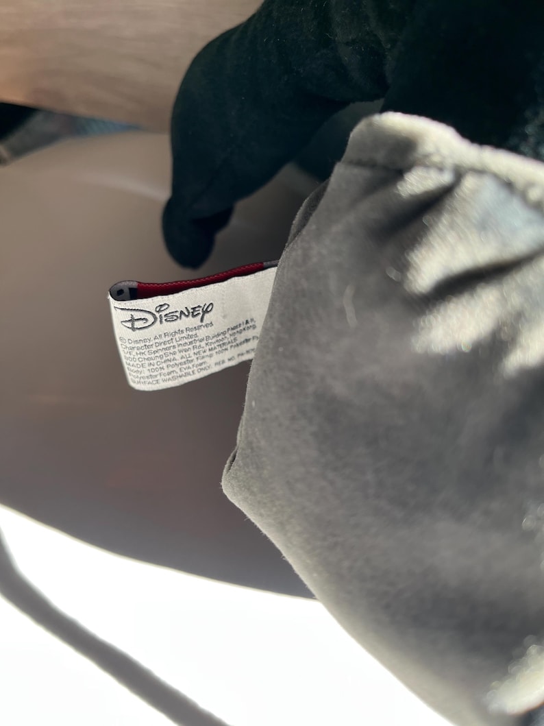 May include: Close-up of a plush toy with a gray body and dark green head. A white tag with the Disney logo and text is visible. The tag is red and white.