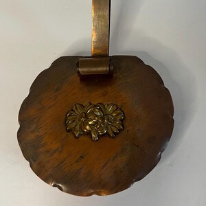 May include: A round, dark brown wooden wall plaque with a scalloped edge and a vertical copper hanging strap. A central brass relief ornament depicts two stylized flowers with textured petals, centered on the polished wood surface.