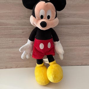 May include: A plush Mickey Mouse toy. The iconic character has a black body, large black ears, and a pink face. He wears red shorts with white buttons, white gloves, and yellow shoes. The toy is standing upright.